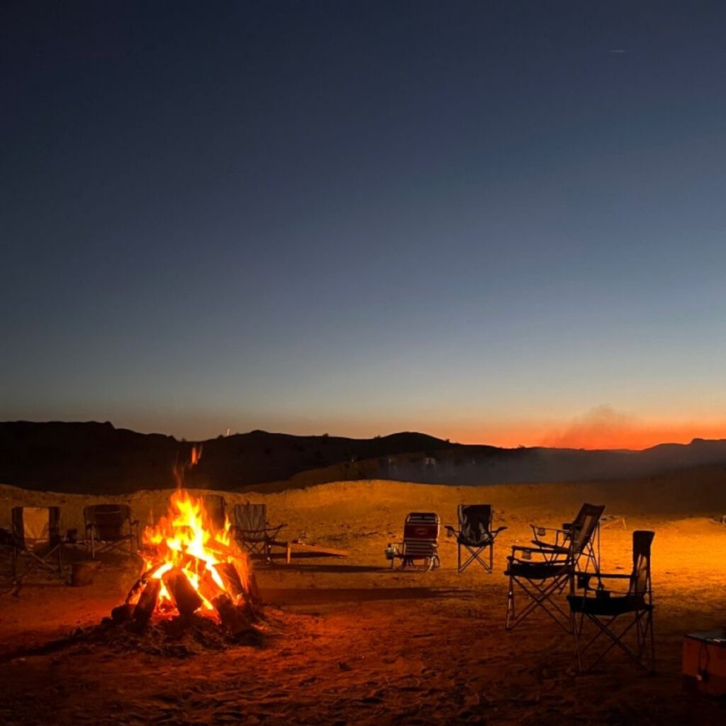Campfire and chairs under sunset desert sky.