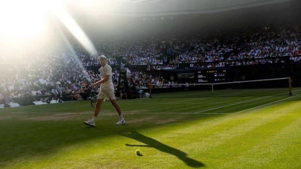 Tennis player walking on grass court in sunlight.