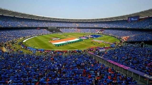Crowded cricket stadium with national flags displayed.