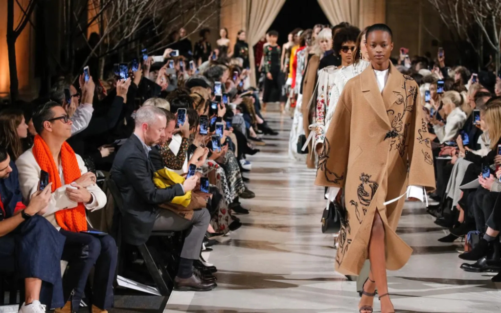 Fashion show runway with audience capturing photos.